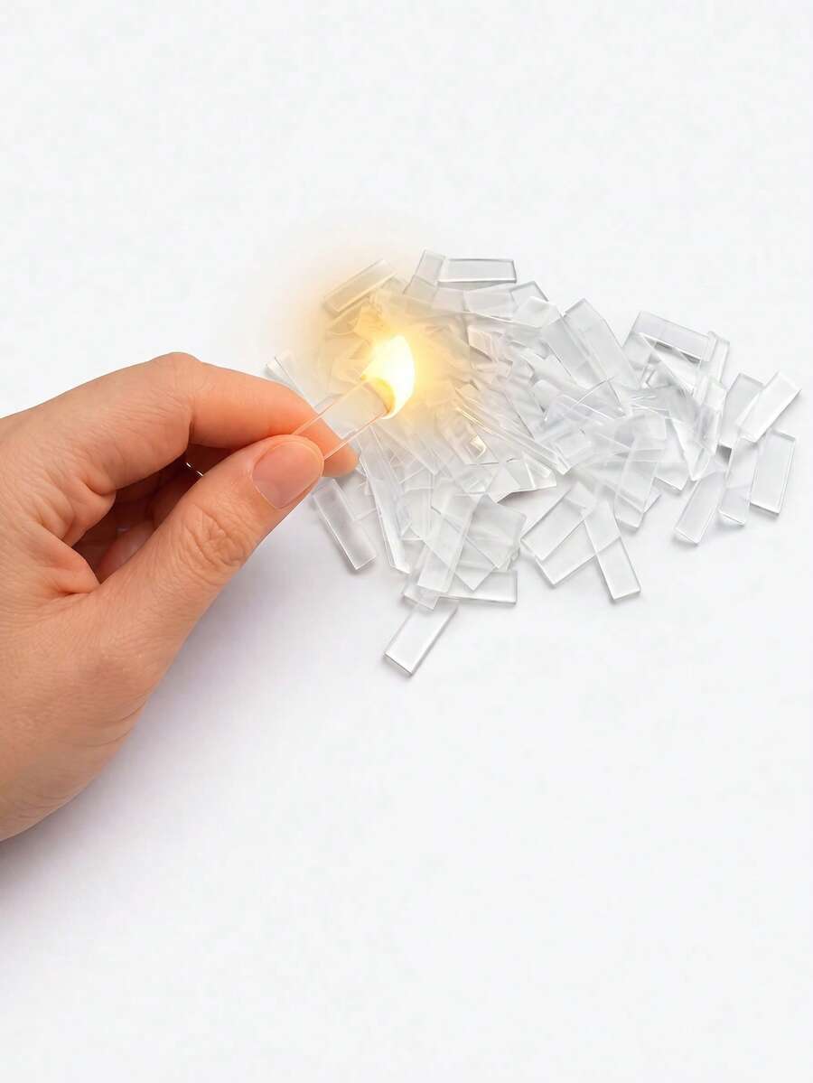 Hand holding a burning clear rectangular fire starter stick next to a pile of unlit sticks on a white background