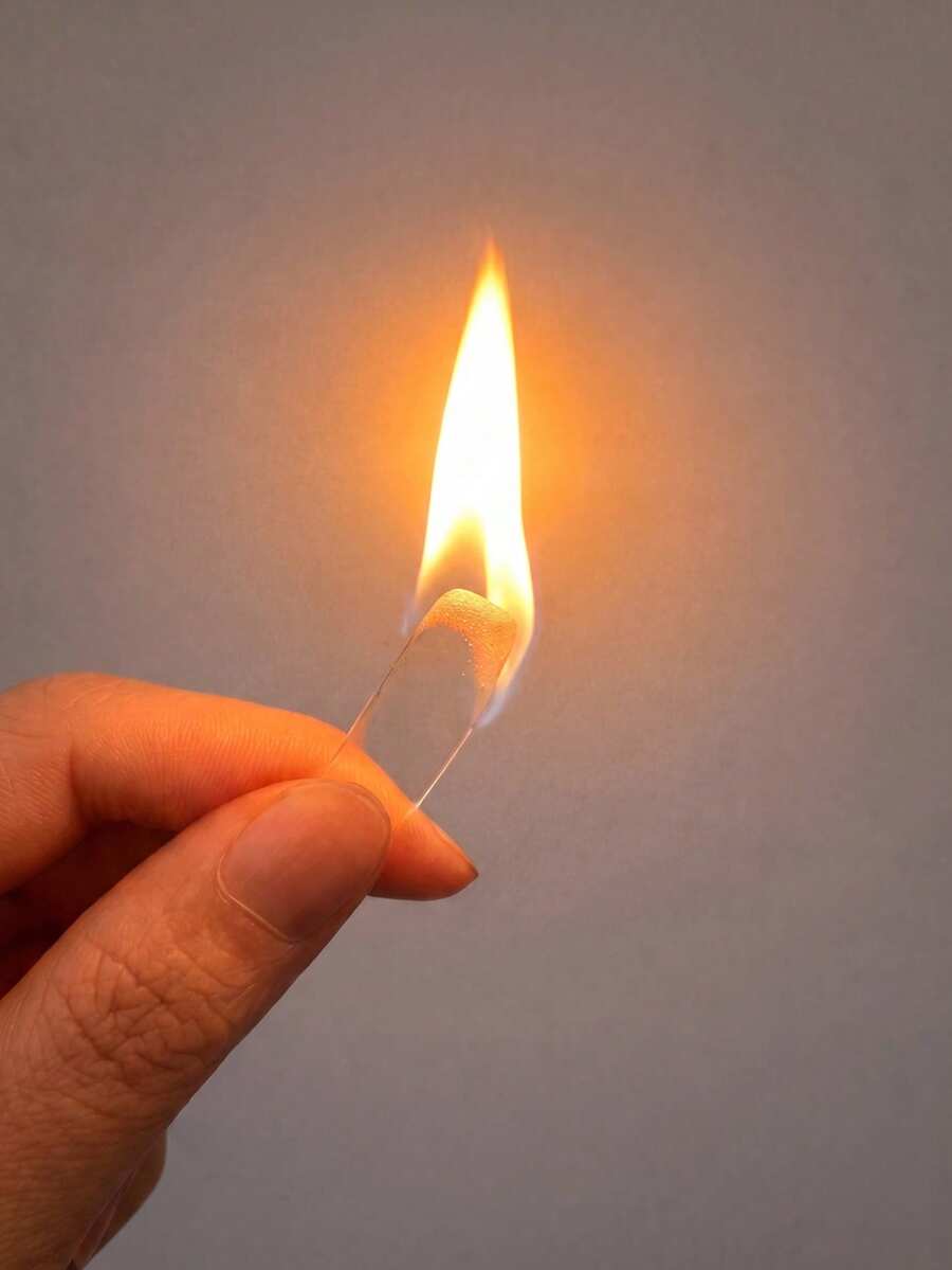 Hand holding a clear rectangular fire starter stick, burning with an orange flame against a grey background
