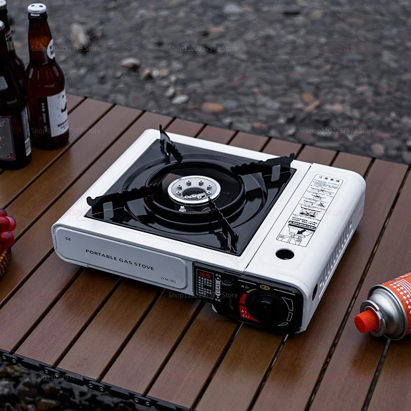 White portable gas stove on a slatted wooden table outdoors, with beer bottles and grapes nearby. PORTABLE GAS STOVE